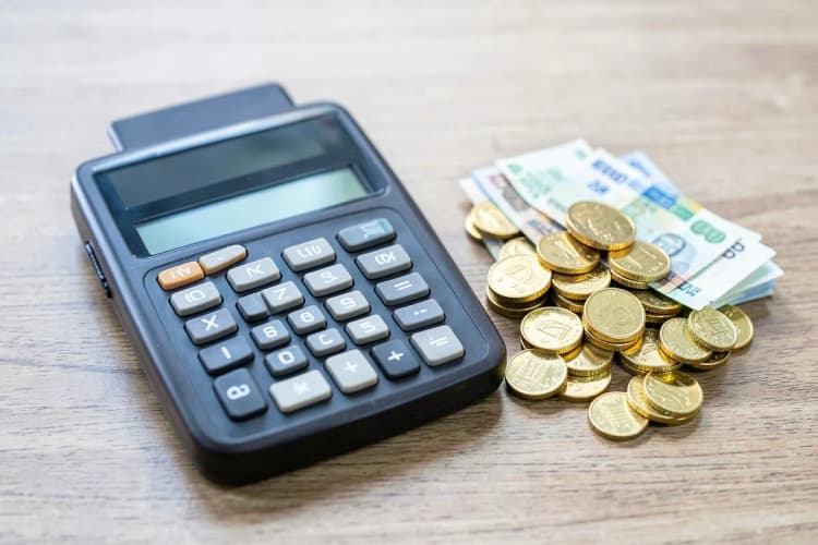 Uma calculadora antiga ao lado de um monte de moedas de ouro e notas de dinheiro, em uma superfície de madeira.