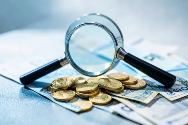 Uma lupa examinando moedas de ouro e notas de dinheiro em uma mesa.
