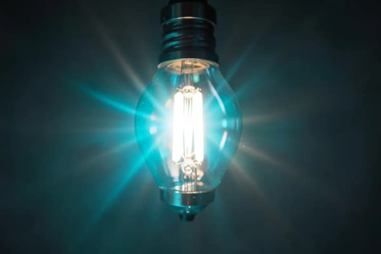 Uma lâmpada incandescente acesa, com raios de luz emanando dela. A iluminação é dramática, com a lâmpada como fonte principal de luz, criando um contraste forte com o fundo escuro. A imagem é um close-up extremo, focando no filamento brilhante da lâmpada.