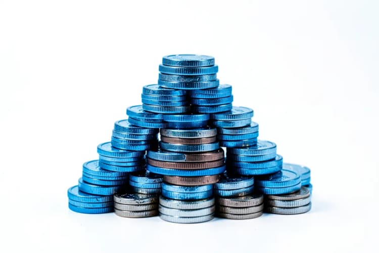 Uma variedade de moedas de diferentes países empilhadas em forma de pirâmide. A iluminação é direcionada, vindo de cima e da esquerda, realçando o brilho metálico das moedas. A imagem é um plano geral, mostrando a totalidade da pilha de moedas.