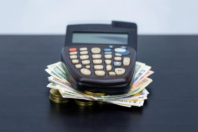 Calculadora antiga sobre uma pilha de moedas de ouro e notas de dinheiro, representando o cálculo de custos.