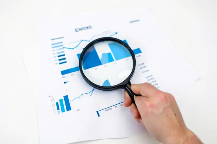Uma mão segurando uma lupa sobre um documento com gráficos e números.