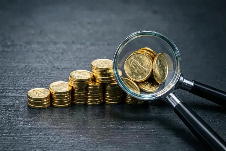Um conjunto de moedas de ouro dispostas em forma de gráfico de barras crescente, com uma lupa focando em uma das barras, sobre uma superfície escura.