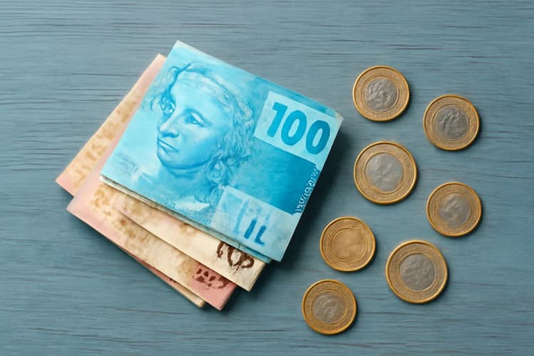 Um conjunto de notas de dinheiro brasileiras dobradas e moedas espalhadas sobre uma superfície de madeira rústica.